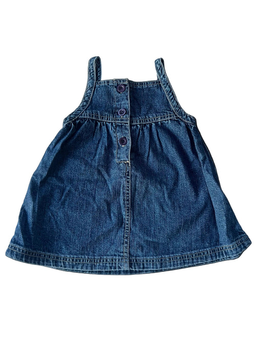 A small blue denim pinafore dress laid flat on a white fabric background.